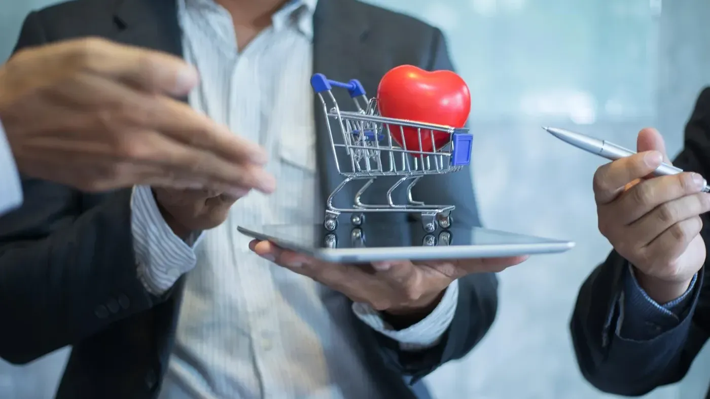 A business professional holding a tablet topped with a miniature shopping cart containing a red heart, discussing e-commerce and customer care with a colleague.
