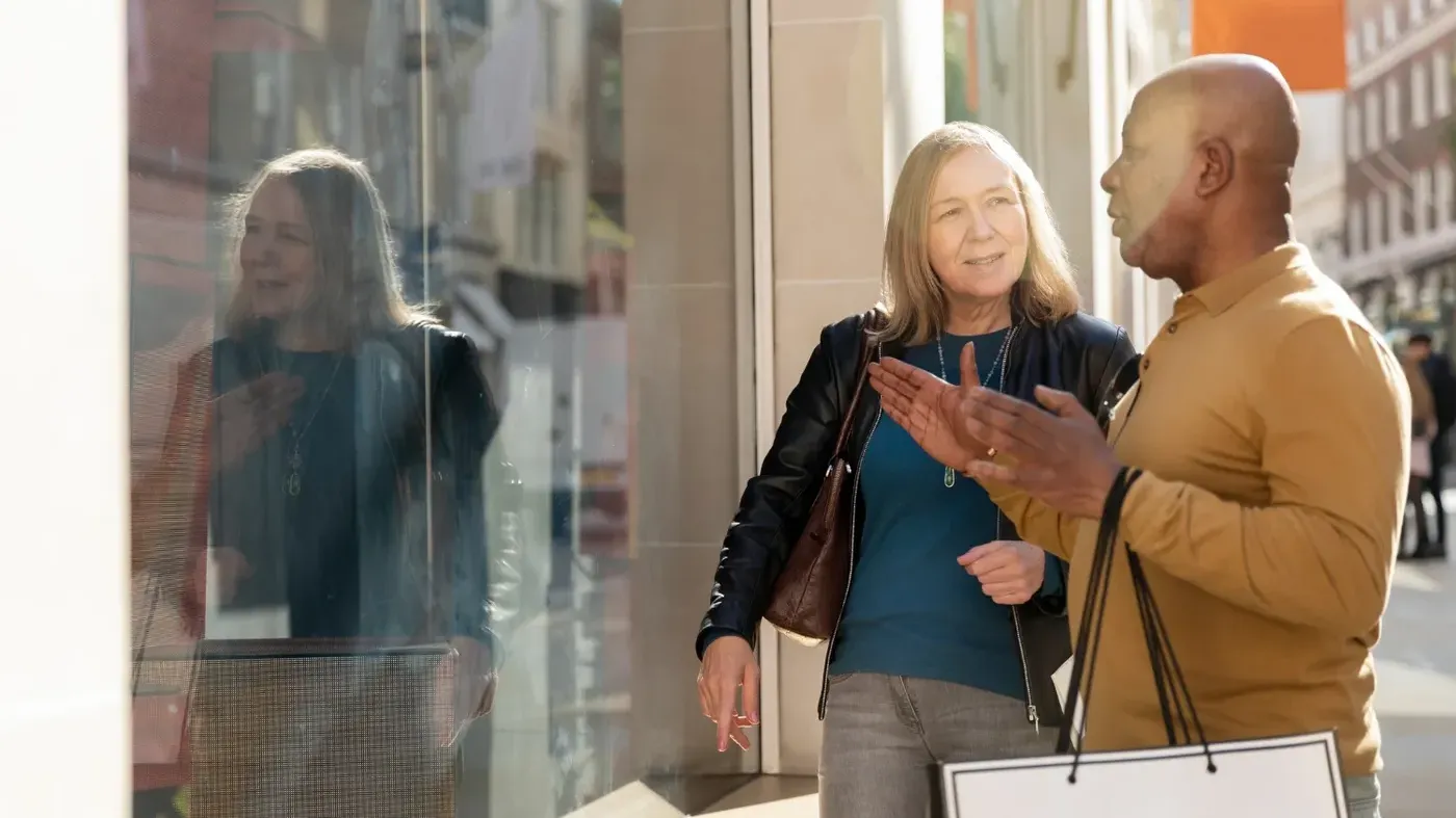 A mature and diverse couple holding shopping bags, stopping to look into a retail store window and discuss products on a sunny city street.