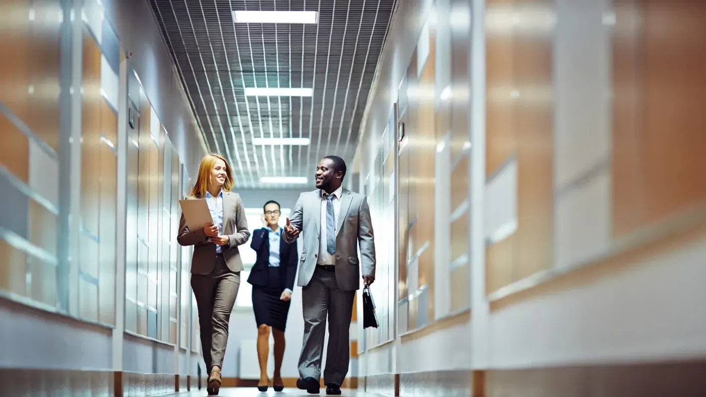 Three smiling corporate executives walking down a modern office hallway while discussing business matters