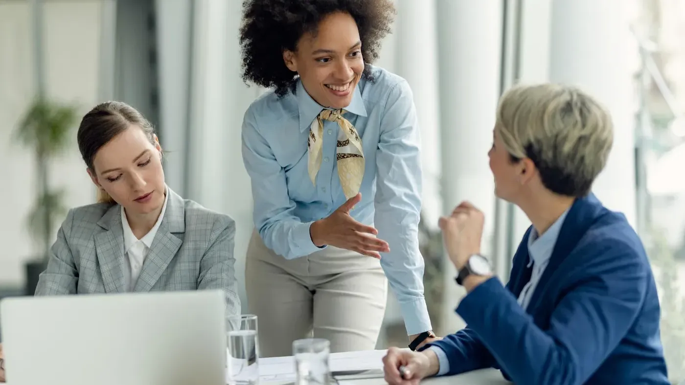A professional woman in a blue blazer speaking to two female colleagues at a desk with a laptop, engaging in a business consultation or training session.