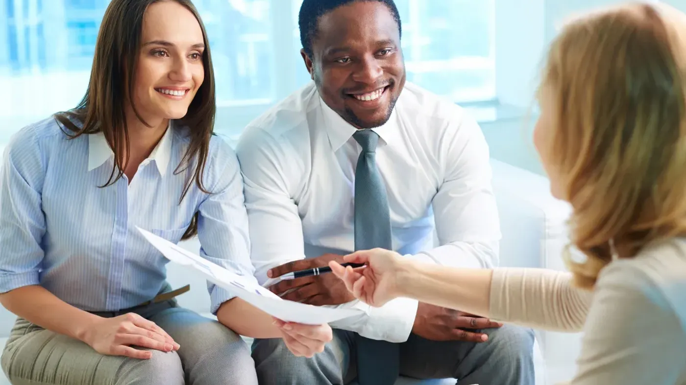 Smiling business partners sitting on a sofa reviewing a document with a female advisor during a professional insurance meeting.