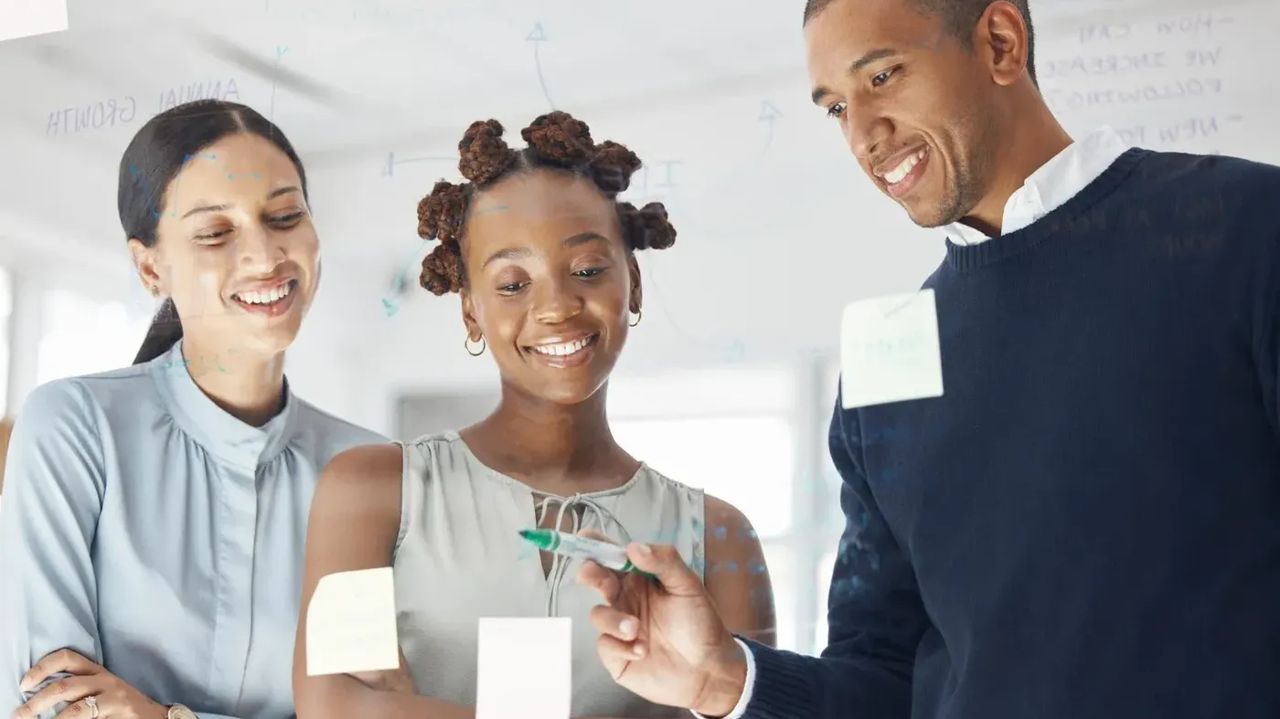 A diverse team of creative professionals smiling and brainstorming ideas using sticky notes and markers on a glass wall in a modern office.