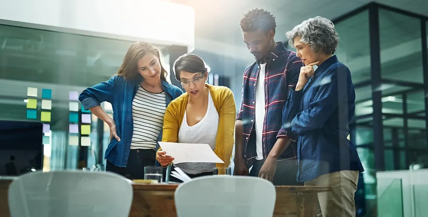 A collaborative team of four diverse colleagues reviewing a document together while standing at a wooden table, viewed through a glass office partition.