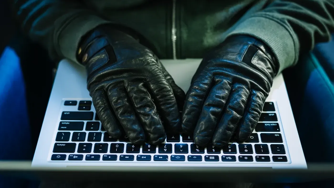 Close-up of a person wearing black leather gloves typing on a laptop keyboard, representing a cybercriminal.