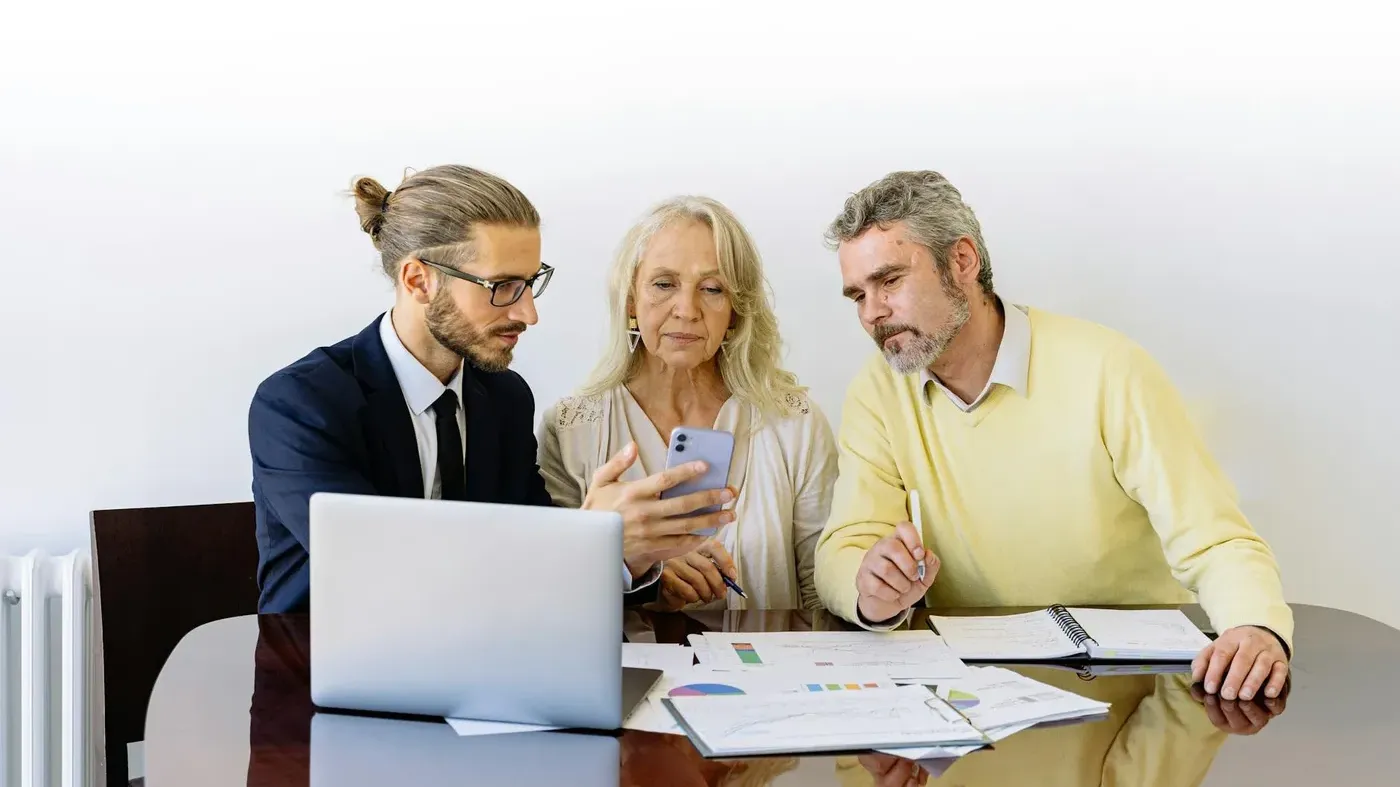 Advisor meeting with two adults at a table reviewing documents and a smartphone beside a laptop, representing insurance planning and financial consultation.