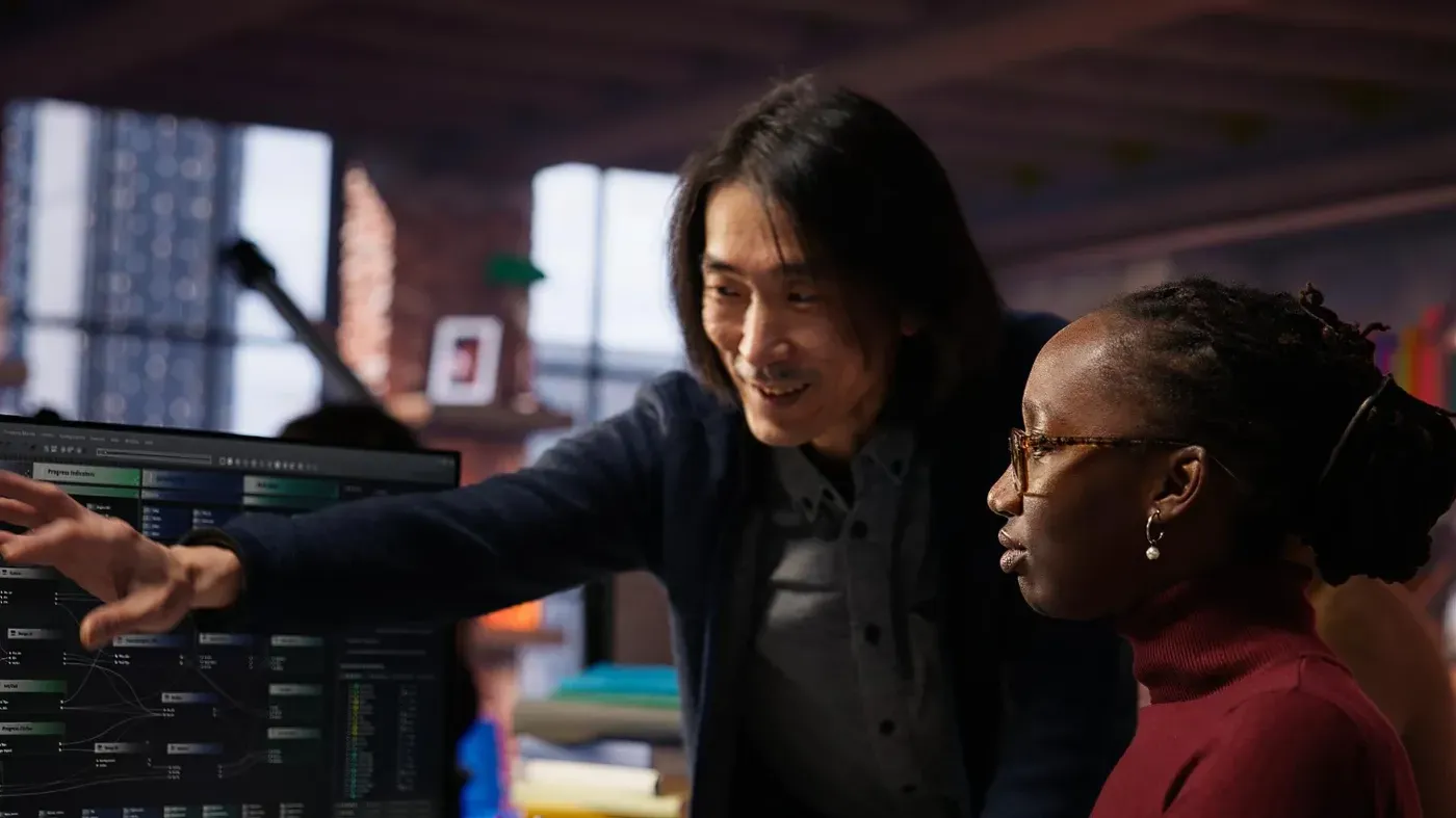 A male professional pointing at a complex digital interface on a monitor while collaborating with a female colleague in a modern office.