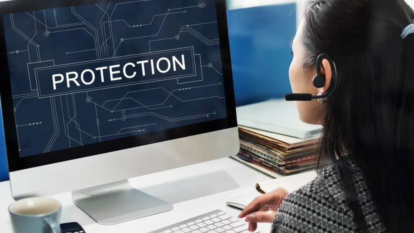 Person wearing a headset working at a desktop computer displaying the word “Protection” over a digital circuit background, representing cybersecurity support and risk protection.