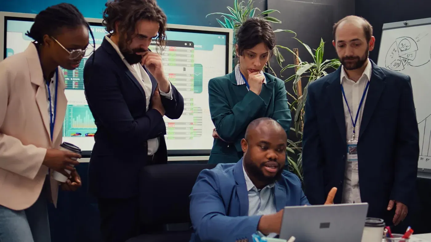 A corporate risk management team analyzing financial data and performance metrics on a laptop and large monitors to mitigate potential liabilities.