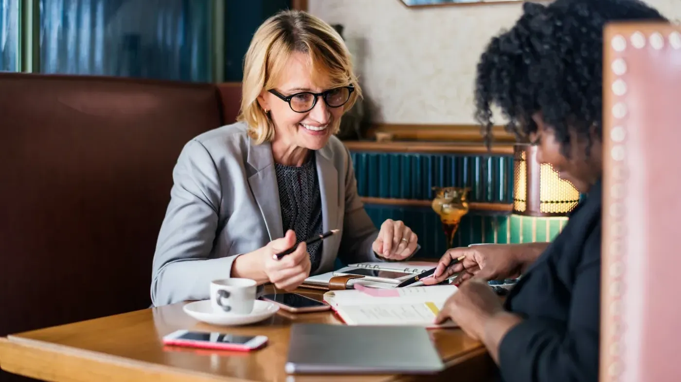 A smiling older female insurance professional in a grey blazer consulting a businesswoman at a wooden table in a cafe setting.
