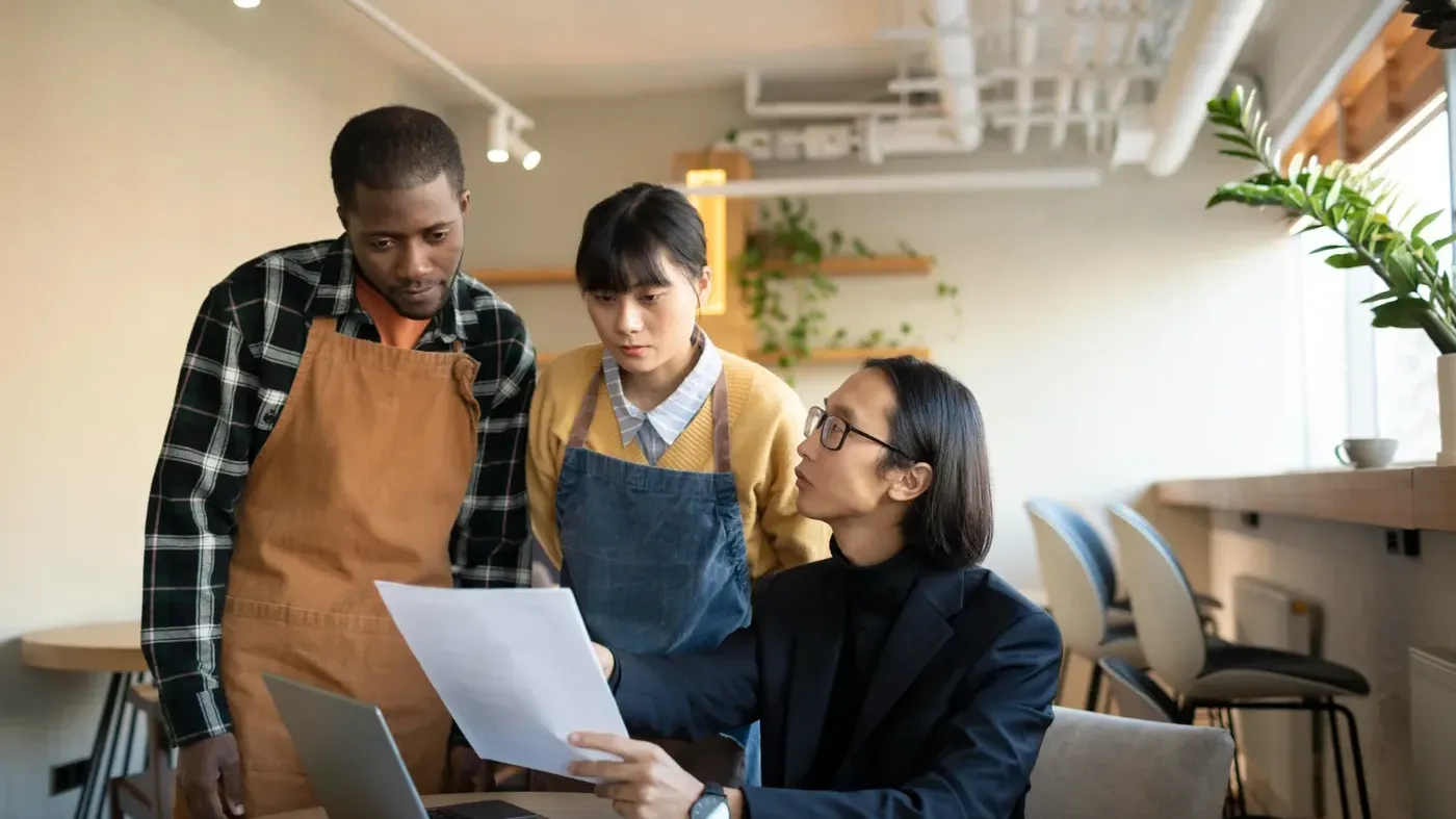 A business advisor in a suit reviewing a document with two restaurant owners wearing aprons during a meeting at a cafe table.