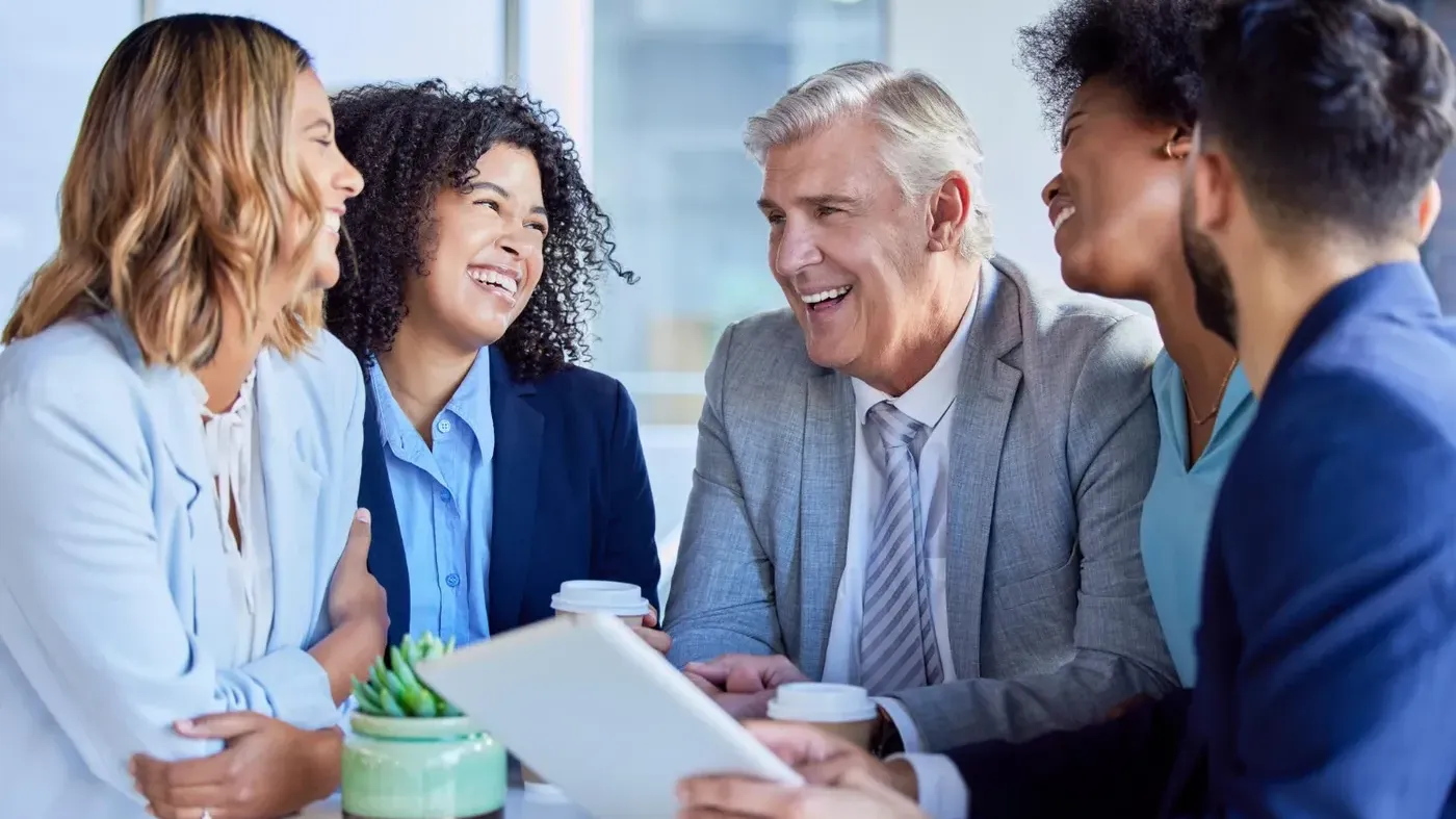 A diverse team of business colleagues laughing and discussing successfully obtaining a liability business insurance quote while gathering around a table with a digital tablet.