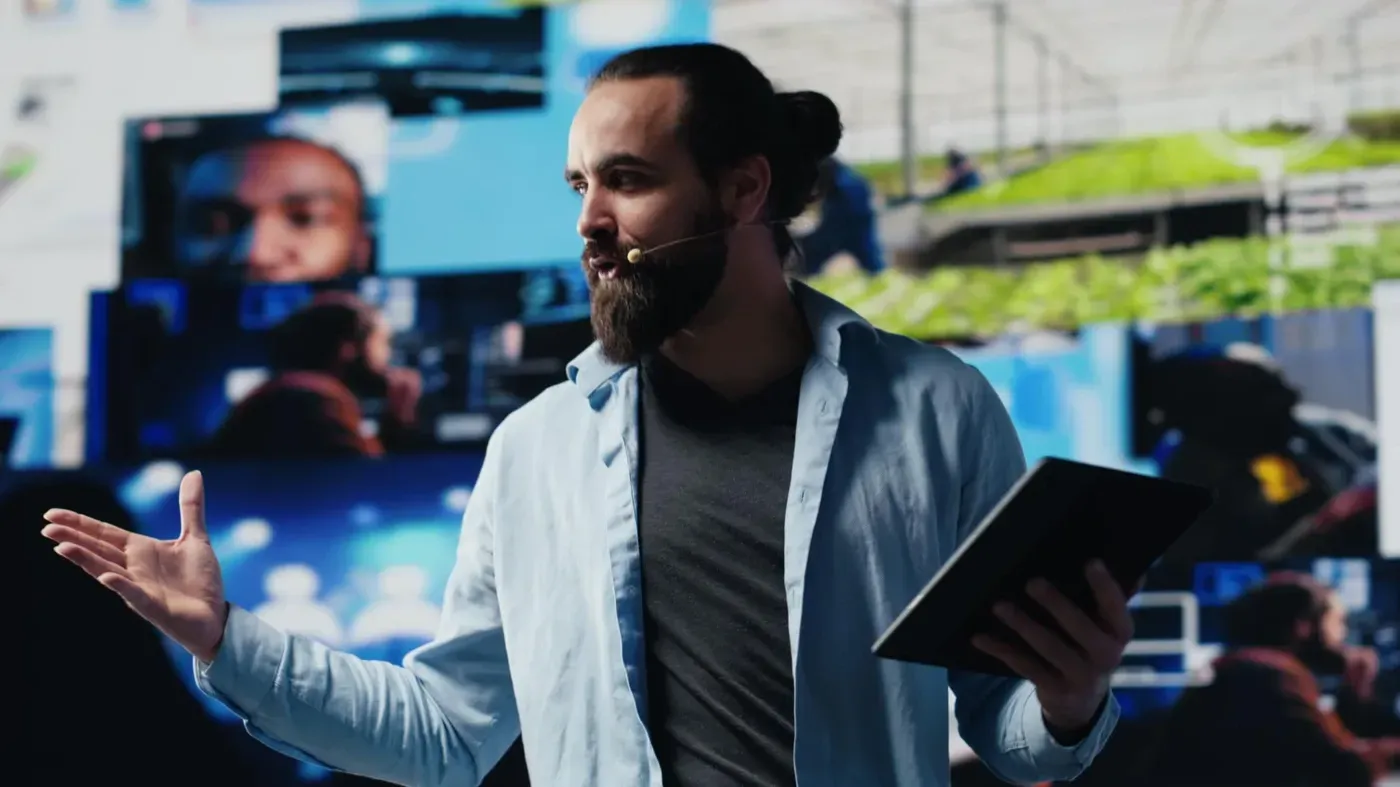 A bearded male presenter wearing a headset microphone holding a tablet and gesturing while speaking in front of a large digital video wall displaying multimedia feeds.