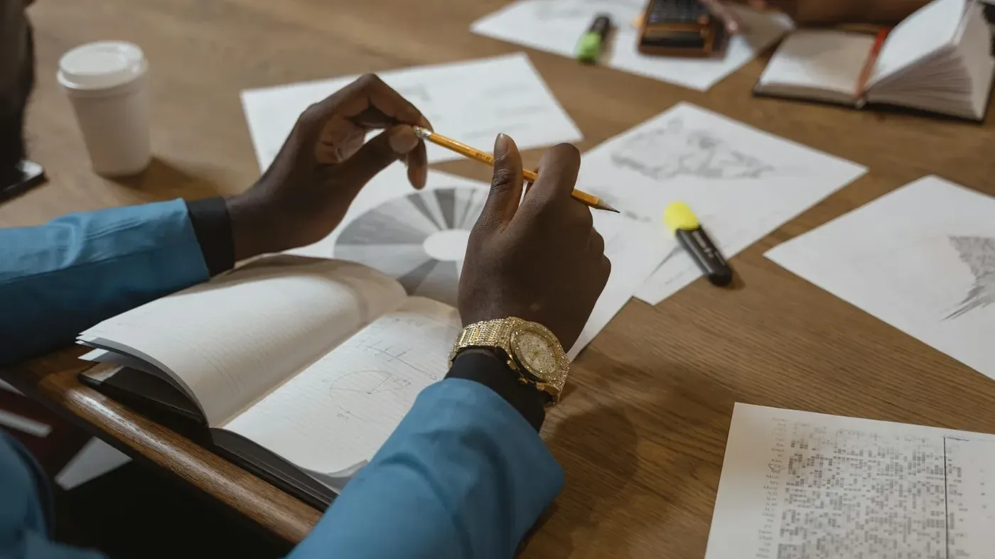 An overhead view of a person’s hands holding a pencil over a notebook, surrounded by financial charts, business charts and graphs on a wooden desk during a business analysis session.