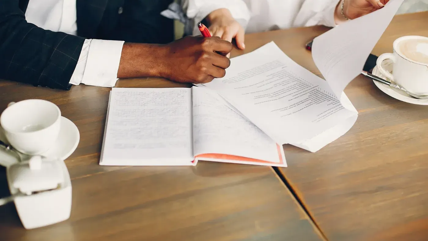 Close-up of a professional signing a document with a red pen at a table with coffee cups, symbolizing the acceptance of a legal agreement or business contract.