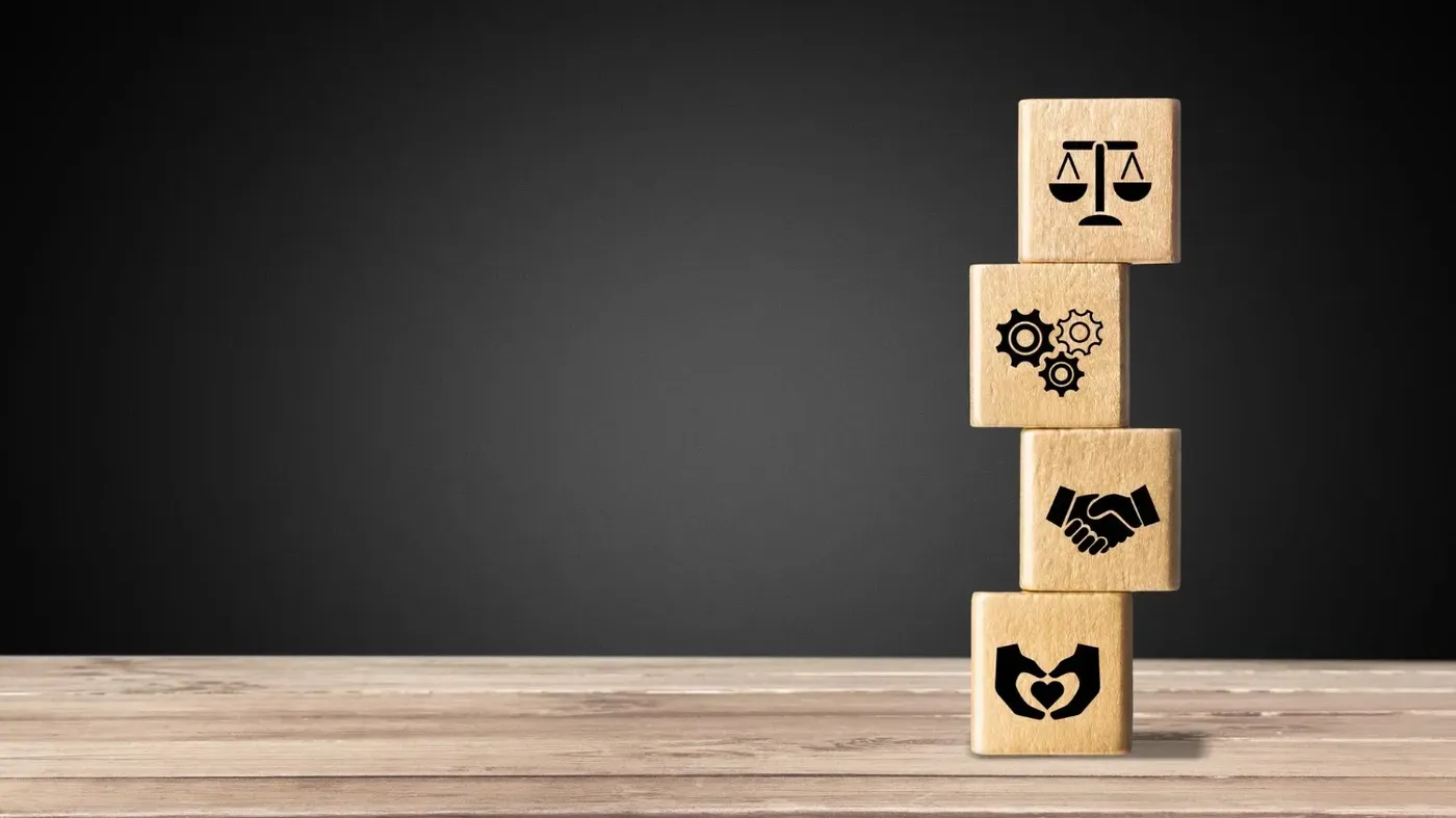 Stacked wooden blocks displaying icons of justice scales, gears, a handshake, and a heart, representing the balance between business operations, ethics, and social responsibility.