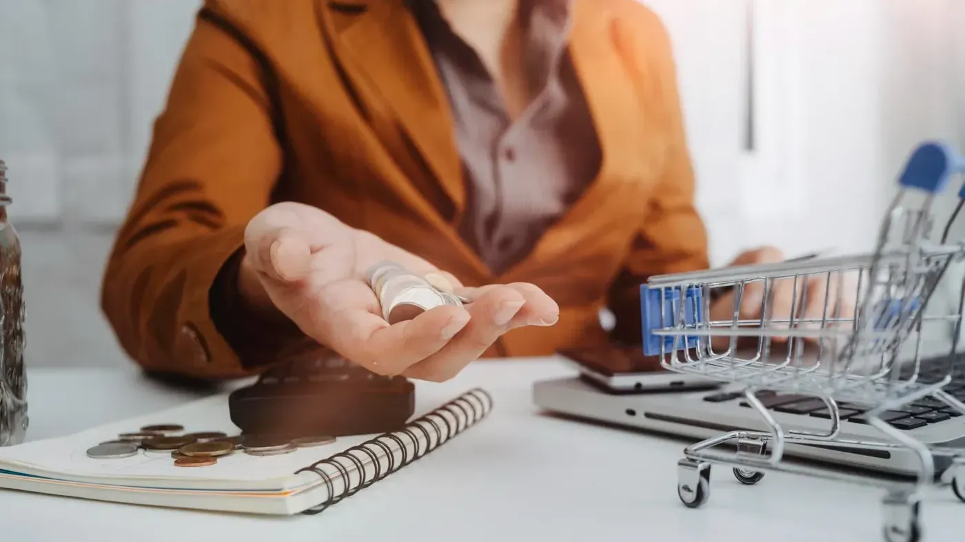 A business woman holding a handful of coins over a white desk containing a laptop, calculator, and miniature shopping cart, representing retail insurance.