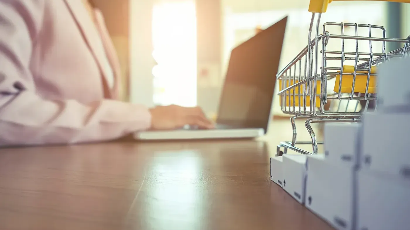 A miniature metal shopping cart and small white shipping boxes sitting on a desk, with a professional working on a laptop in the blurred background.