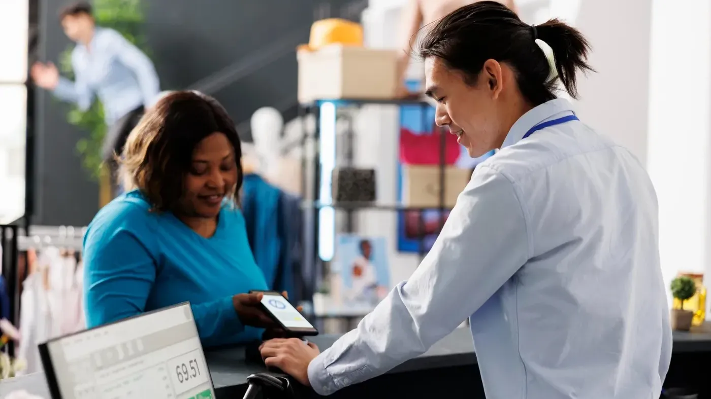 A male cashier holding a payment terminal while a female customer completes a contactless payment using her smartphone at a retail checkout counter.