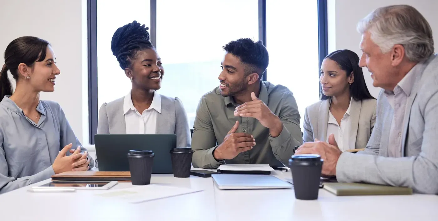A group of diverse professionals including stakeholders, management, IT staff, and employees engaging in a strategic planning discussion at a white conference table with coffee cups and notebooks.