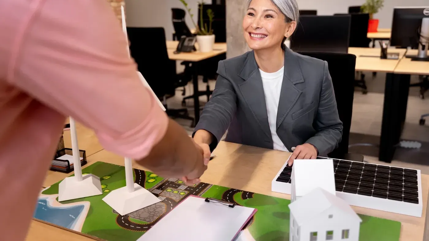 A smiling small businesswoman with grey hair shaking hands with an insurance broker over a desk.