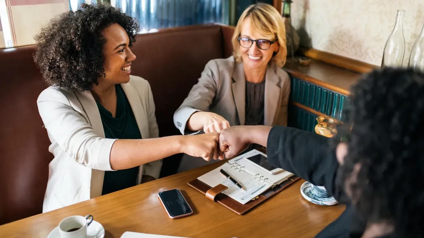 Three diverse professional women celebrating teamwork with a group fist bump while sitting at a table with notebooks and coffee.