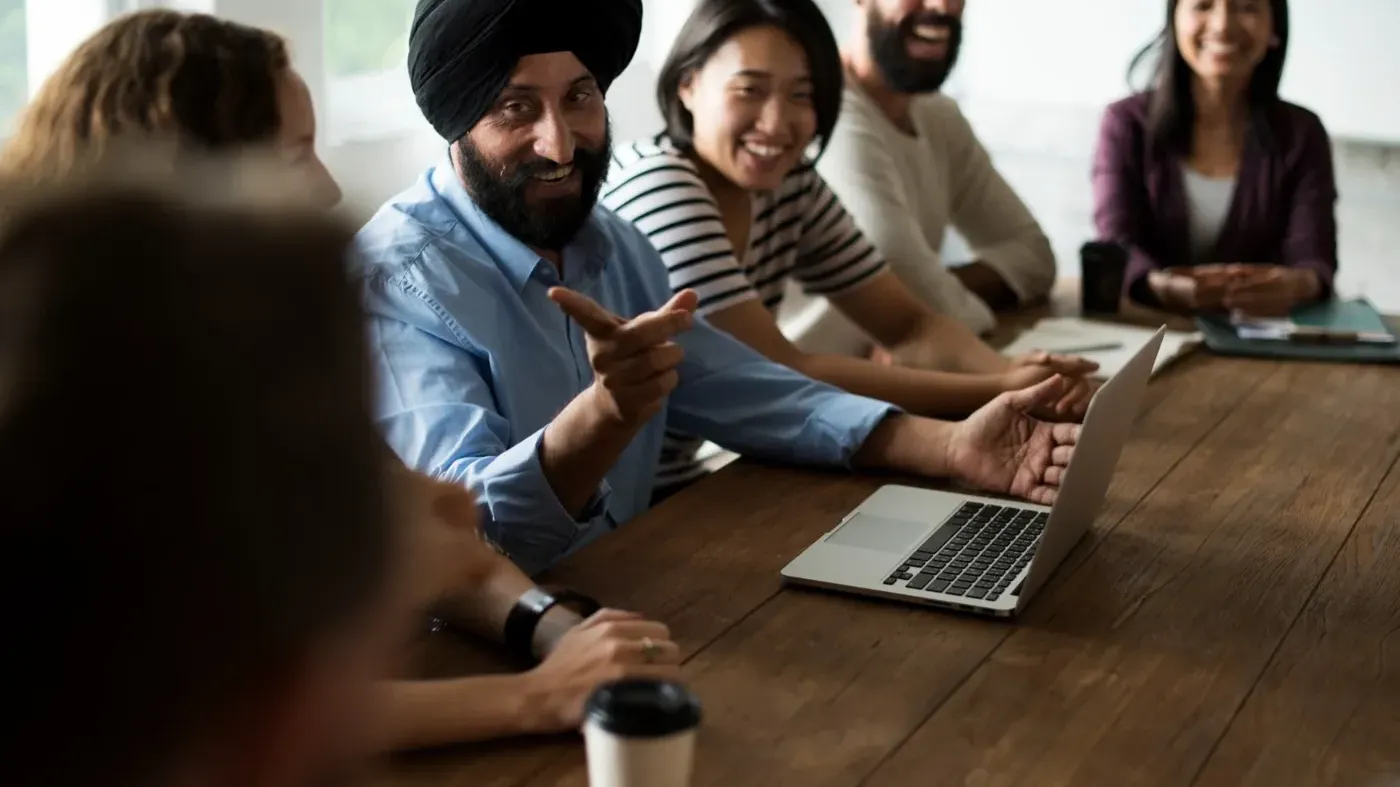A smiling business professional wearing a black turban gesturing with his hand while speaking to his diverse small business team during a meeting at a wooden table.