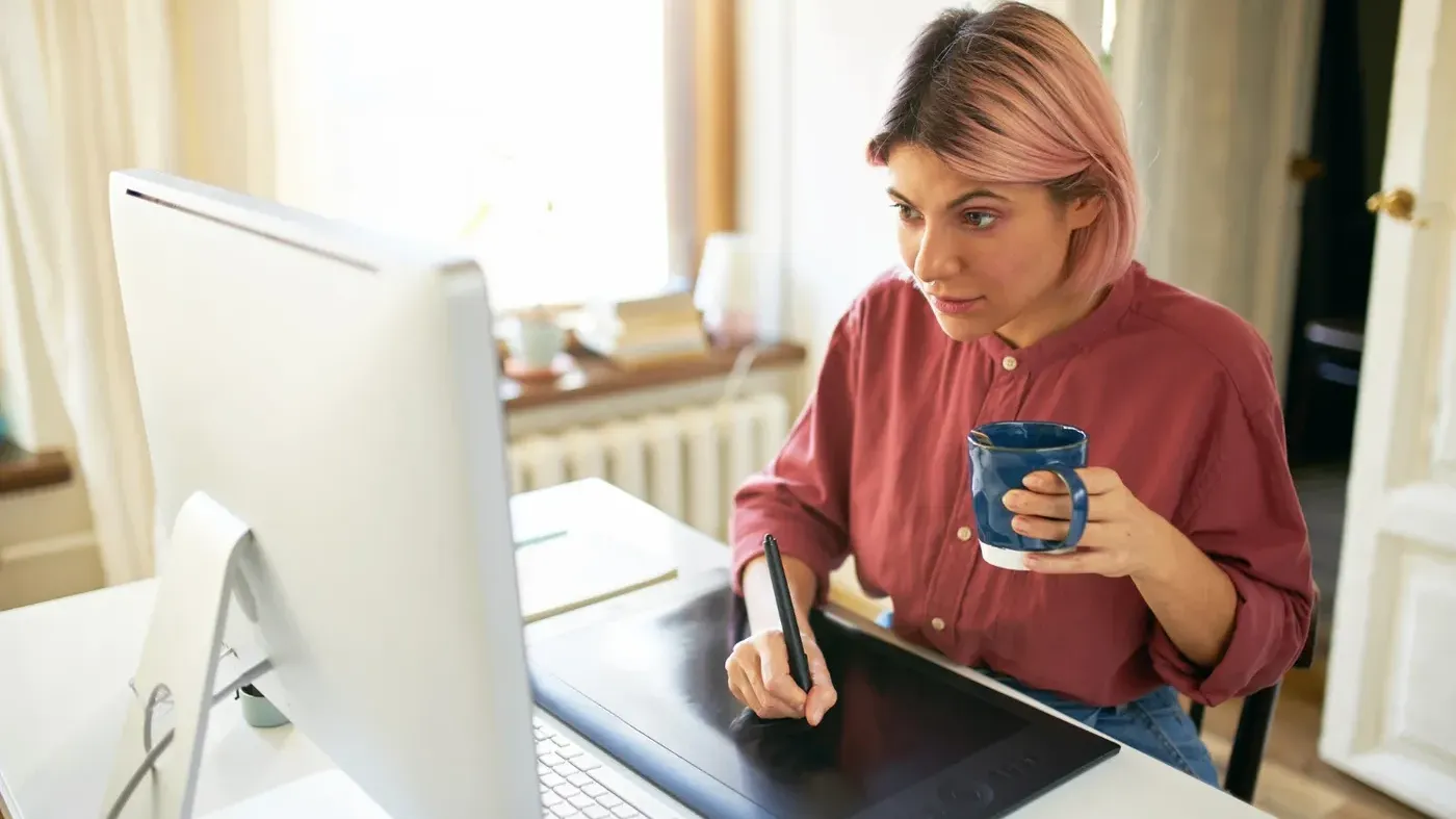 A creative professional with pink hair holding a stylus and a blue mug while working on a digital drawing tablet in front of a large computer monitor.