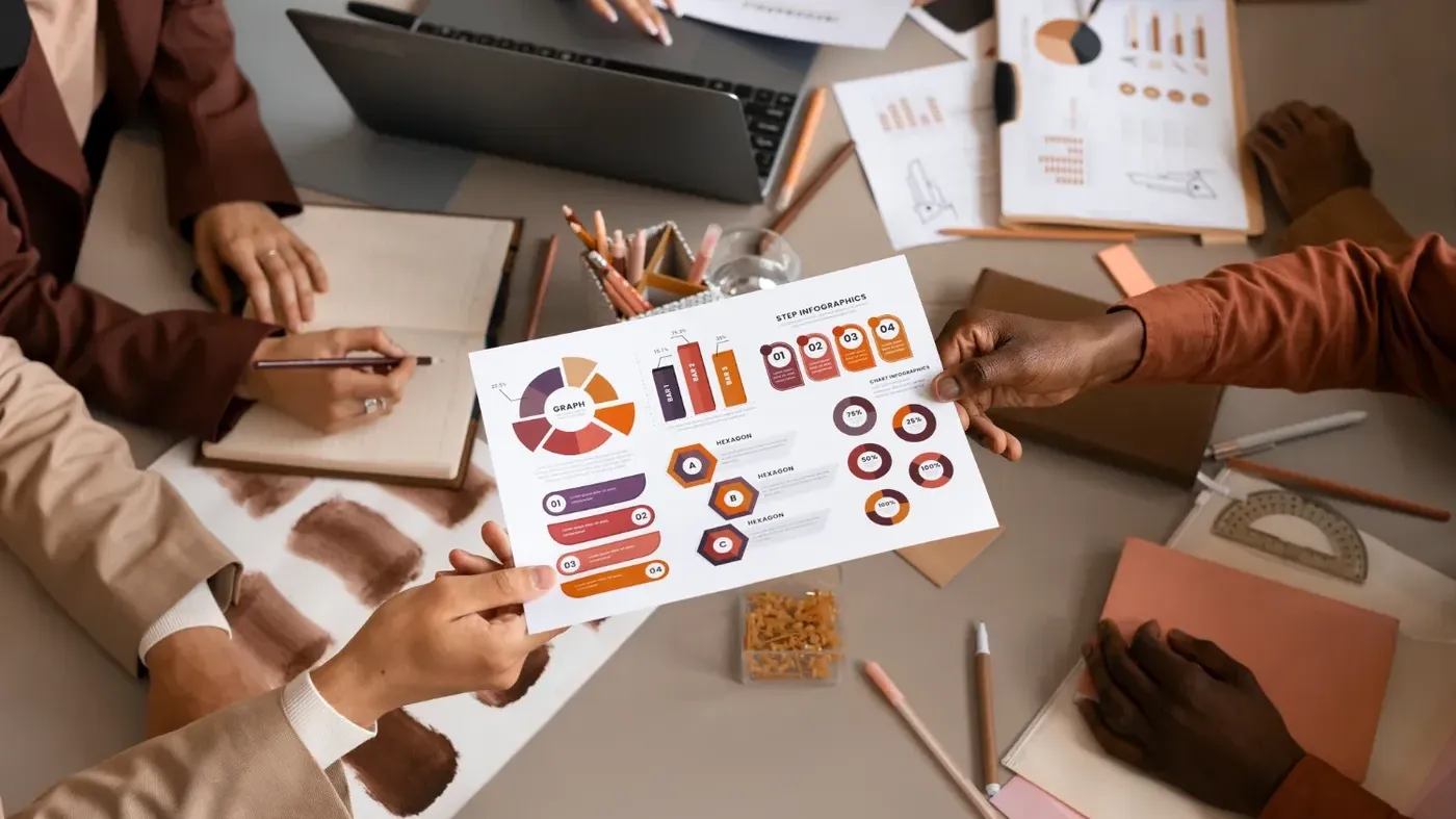 An overhead view of a business team collaborating at a table, exchanging a document featuring colorful charts and infographics amidst laptops and notebooks.