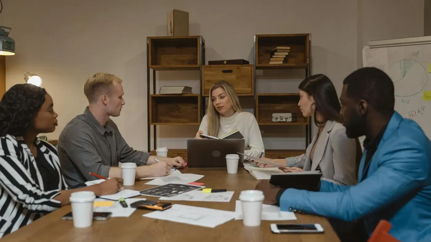A female business leader in a white sweater holding a notebook and speaking to her diverse team during a discussion at a conference table.
