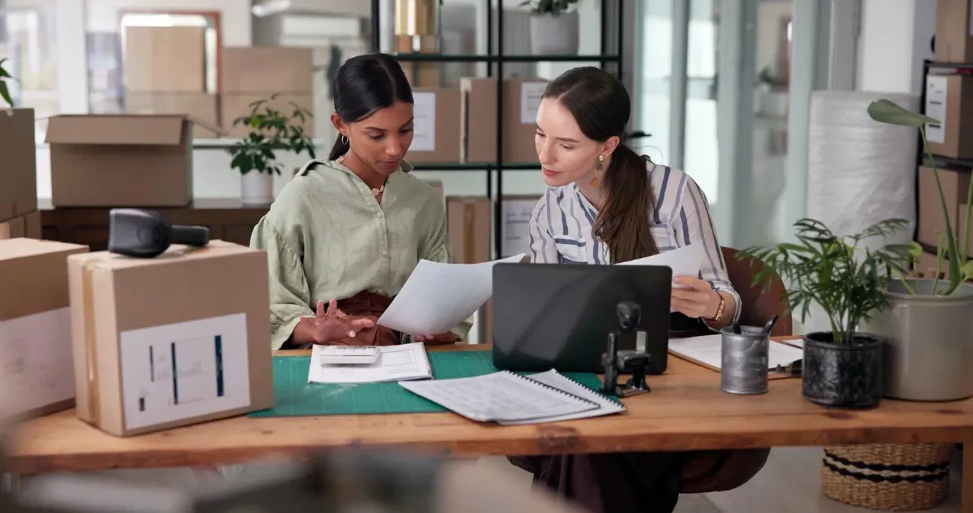 Two female small business owners reviewing insurance coverage and shipping manifests at a wooden desk surrounded by cardboard inventory boxes.
